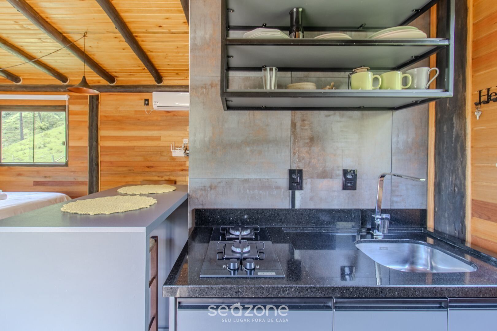 A modern kitchen with a granite countertop, stainless steel sink, and a built-in oven. There is a shelf above the sink holding dishes and a hanging pot rack.