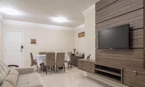 A living room with a sofa, dining table, chairs, a TV mounted on a wooden wall, and a cabinet. - 3rooms apt w Suite and bbc Close to Beaches Egv302 (Balneário Camboriú)