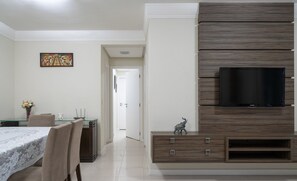 Ein modernes Wohnzimmer mit Flachbildfernseher an einer Wand aus Holzpaneelen, einem Essbereich mit Glastisch und Stühlen sowie einem Flur, der in einen weiteren Raum führt.