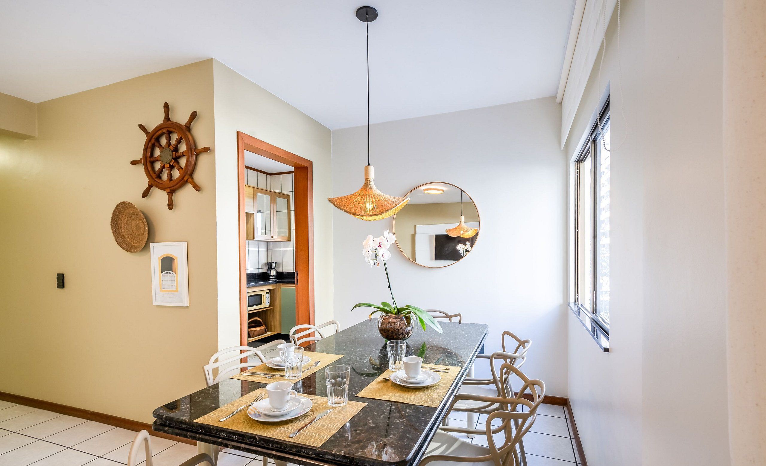 A dining area with a marble table, white chairs, a ship's wheel decoration, a hanging light fixture, and a mirror on the wall.