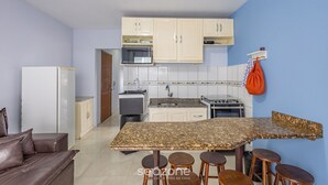 A compact kitchen with white cabinets, a microwave, a stove, and a sink. There is a dining table with stools and a brown leather couch in the living area. - Beachfront Apartment in BC Great Location Eit302 (Balneário Camboriú)