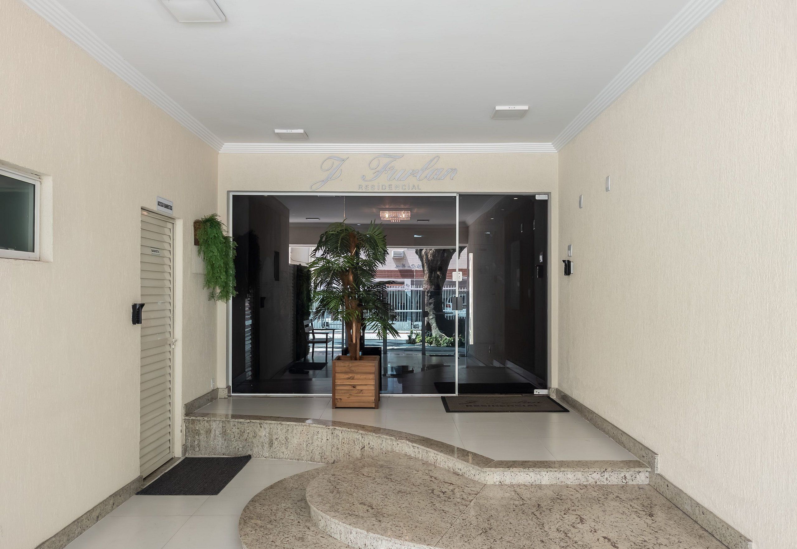 A modern hotel entrance with a reception desk, a potted plant, and a glass door leading to the interior.