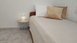 Una cama con almohada blanca y color crema, una mesita redonda pequeña con una lámpara blanca y una cesta de alambre.