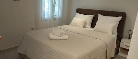 A bedroom with a bed, pillows, a nightstand, a bedside table, and a window with curtains.