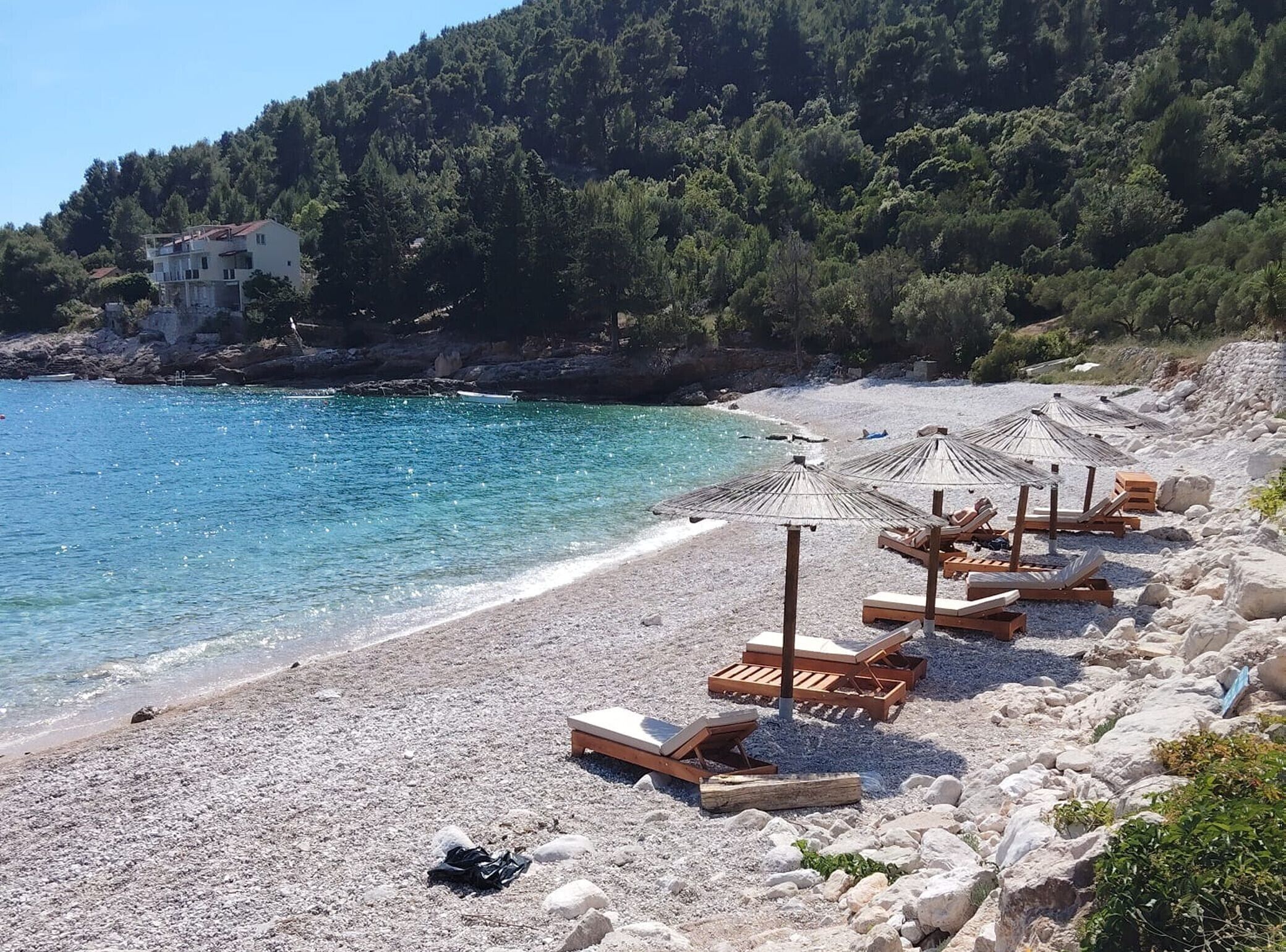 Una playa de guijarros con tumbonas y sombrillas de madera, un mar azul cristalino y un promontorio boscoso al fondo.