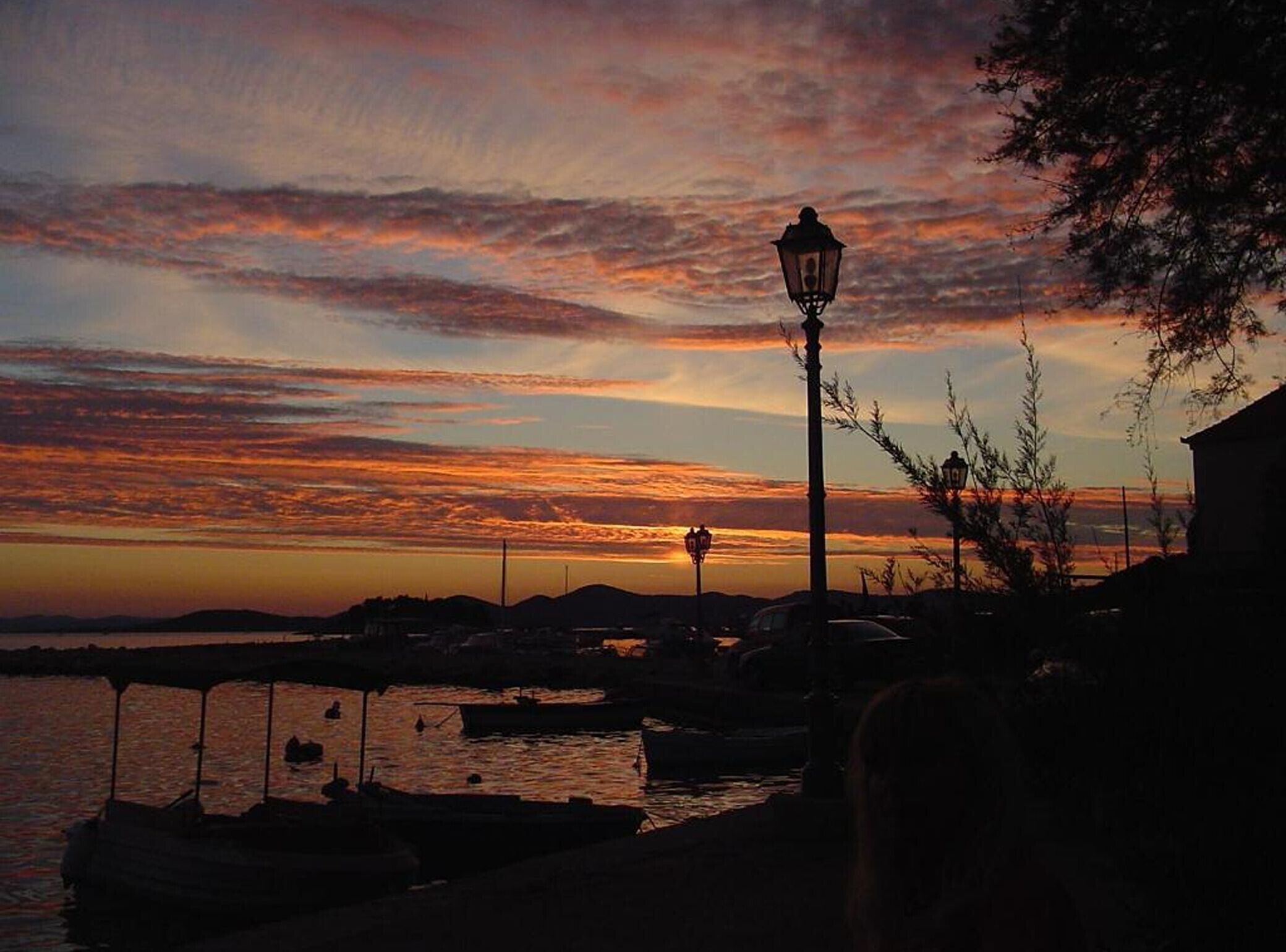 Un atardecer sobre un puerto con barcos, un farol y un edificio al frente.