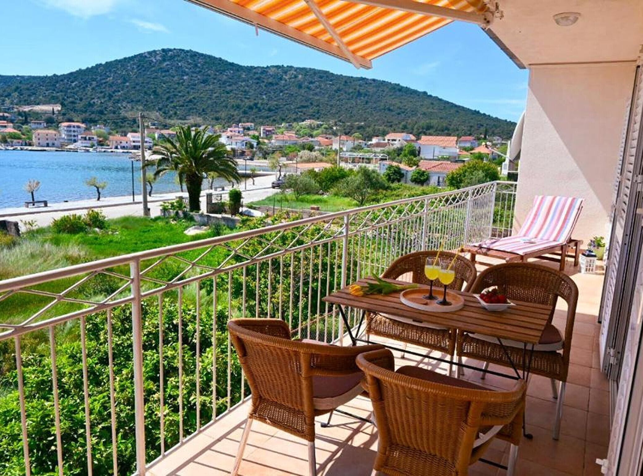A balcony with wicker furniture, a table with drinks, and a view of a coastal town and hills.