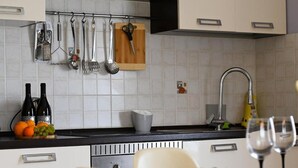 Cocina moderna con armarios blancos, encimera negra y diversos utensilios colgando. Hay un lavabo con grifo, una botella de vino y un vaso sobre la encimera.