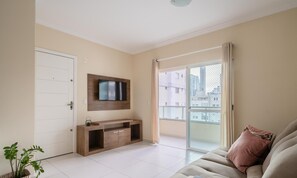 Un salon avec un téléviseur fixé au mur, une porte-fenêtre coulissante donnant sur un balcon et un canapé beige.