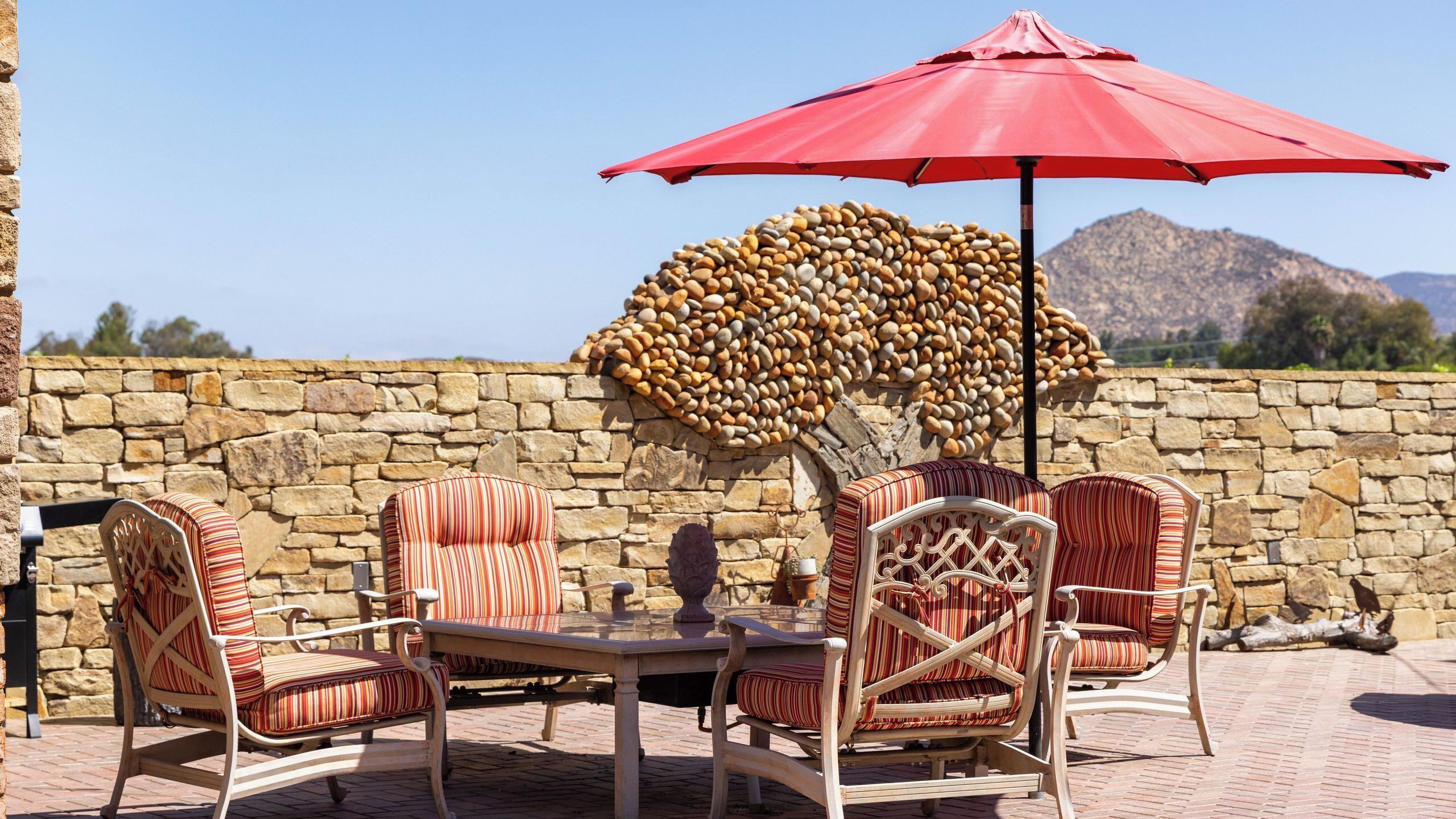 An outdoor seating area with wicker chairs and a table, a red umbrella, and a stone wall.