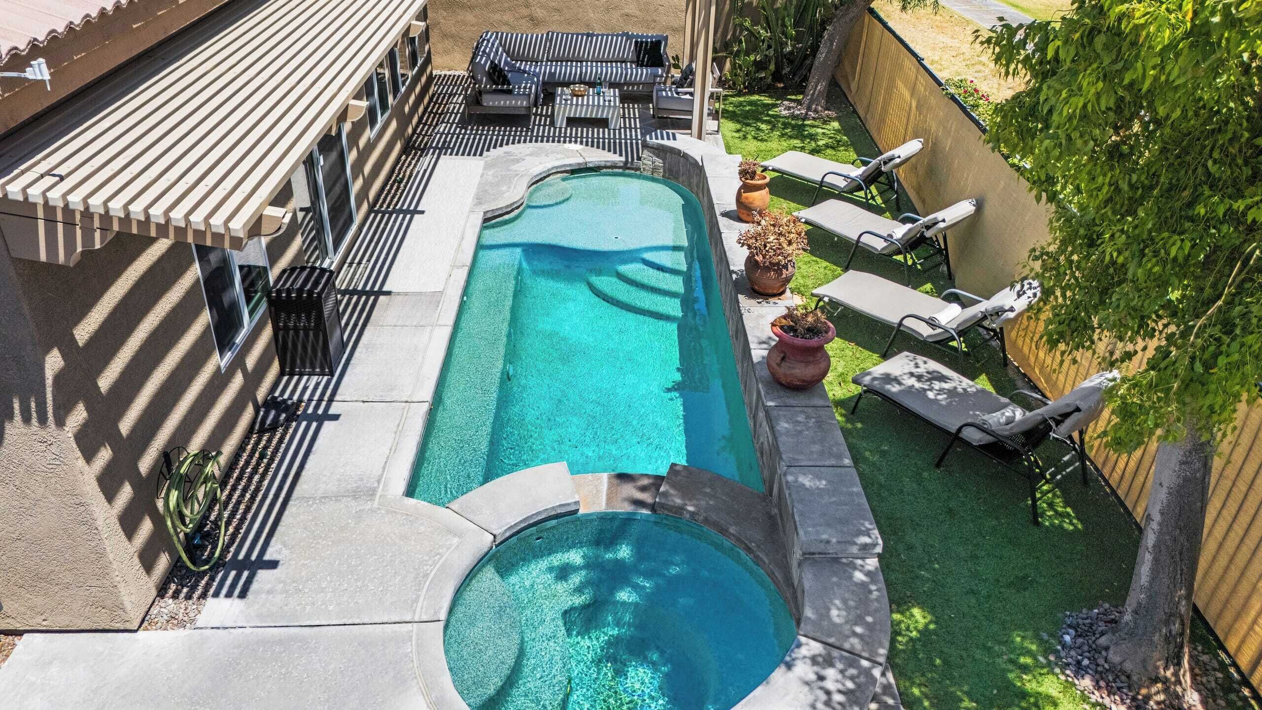 Une piscine rectangulaire avec un petit jacuzzi, entourée d’une terrasse dotée de chaises longues et d’une pergola.