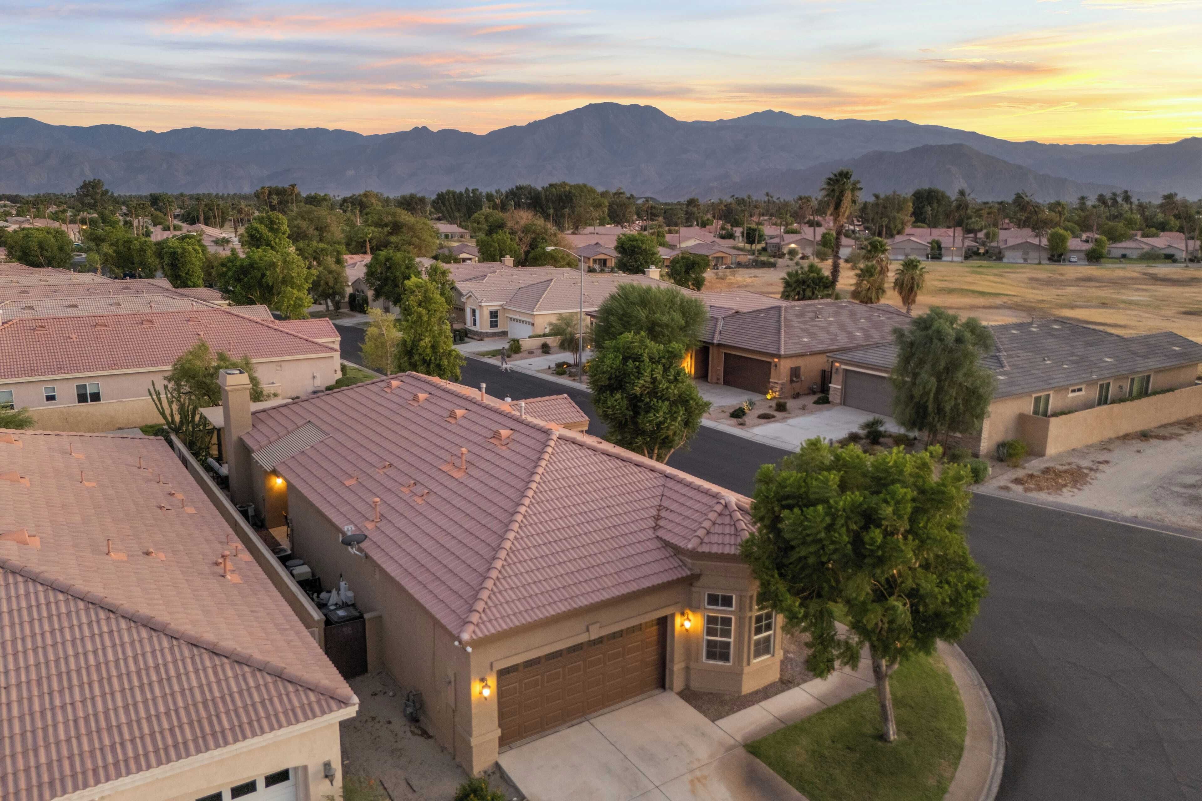 A residential neighborhood with houses featuring red-tiled roofs, surrounded by greenery and mountains in the background.