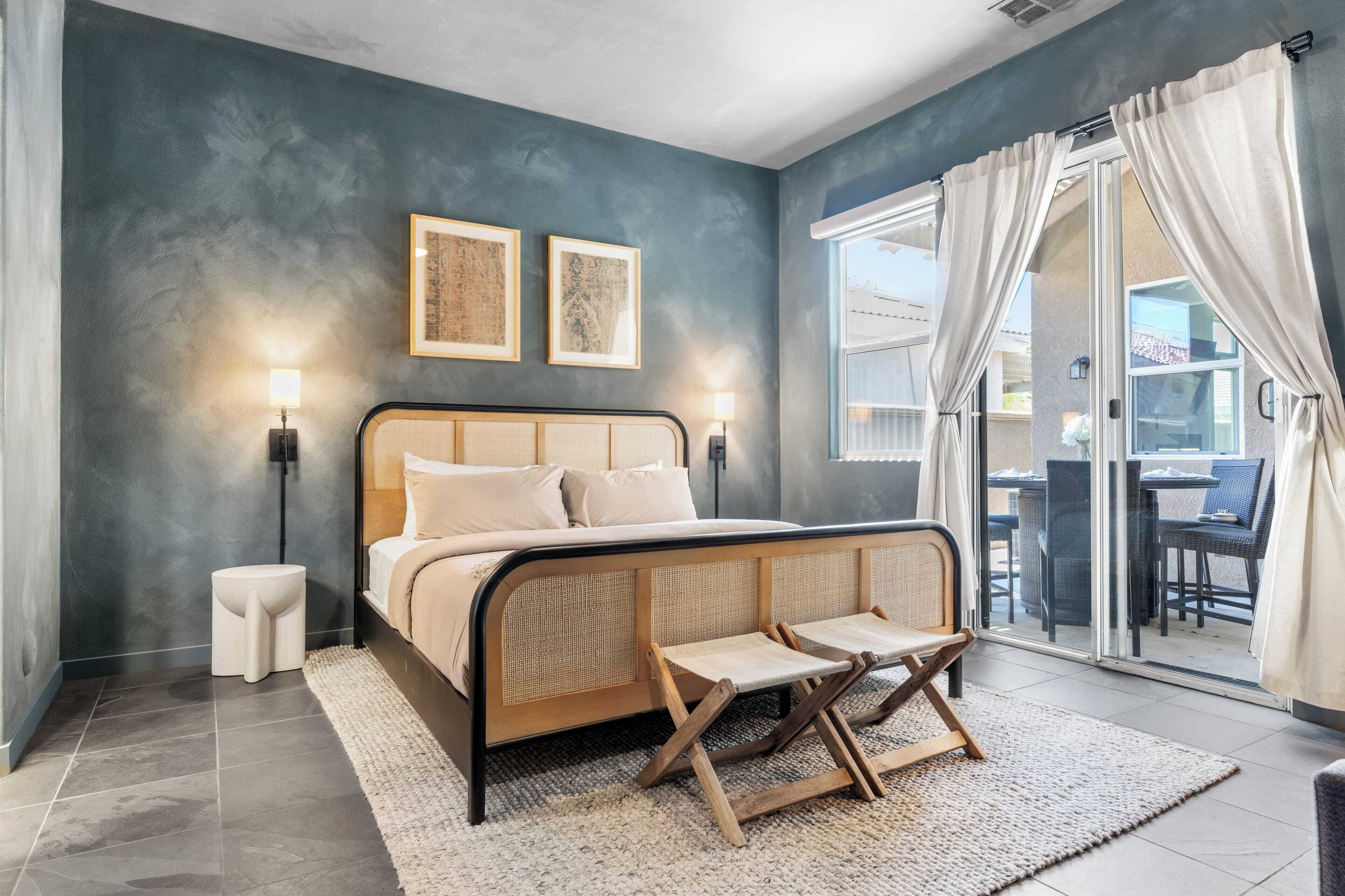 A bedroom with a bed, bedside tables, a small stool, a rattan chair, and a view through a glass door.