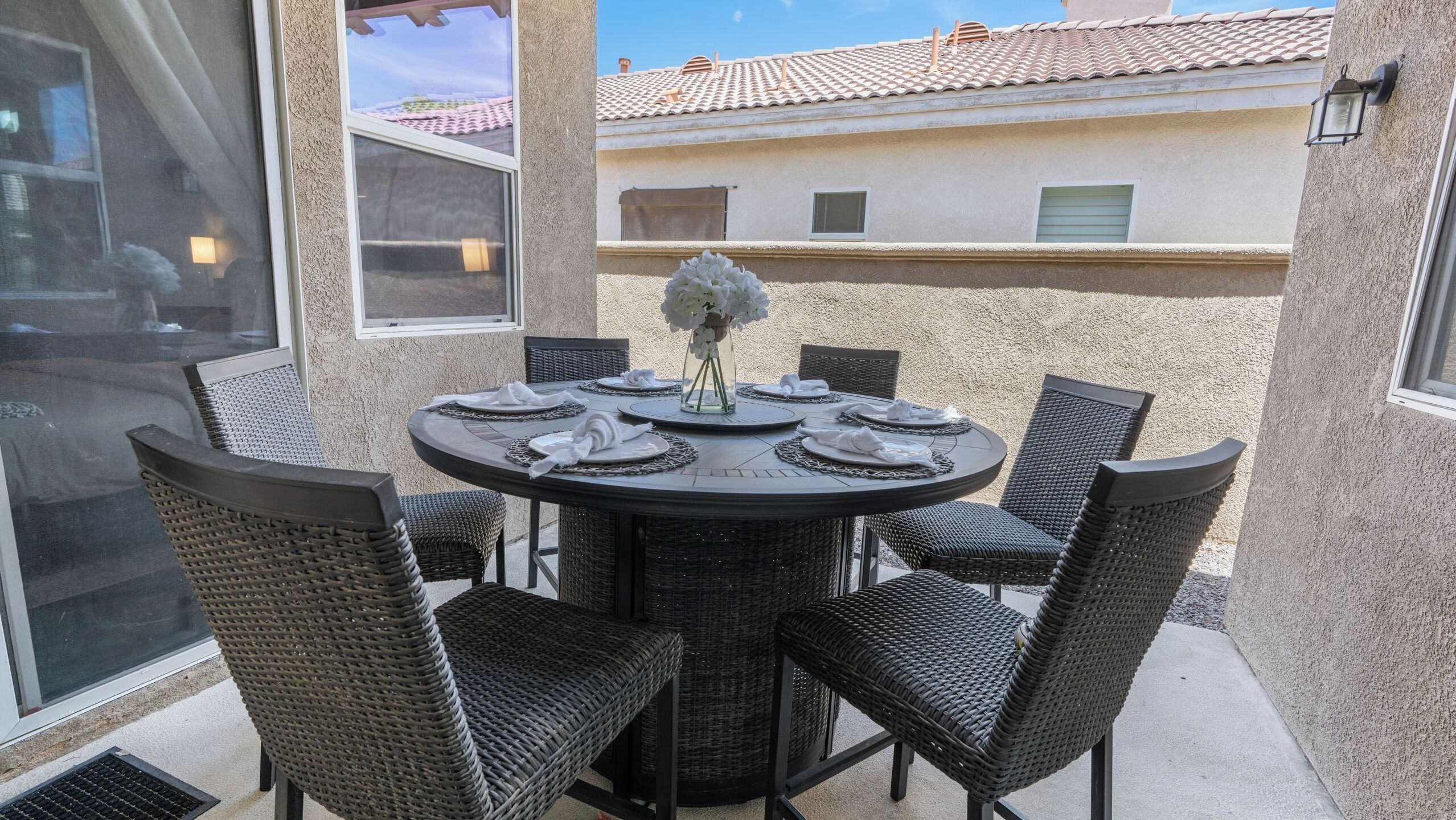 A round outdoor dining table with wicker chairs, a vase with white flowers, and a clear view of a building with a tiled roof in the background.