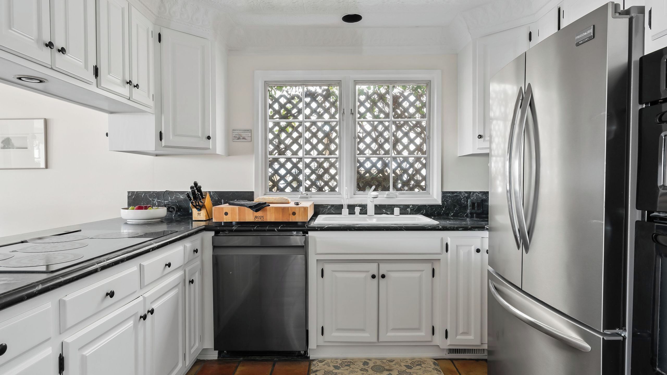 A kitchen with white cabinets, a stainless steel refrigerator, a sink, and a window with a lattice screen.