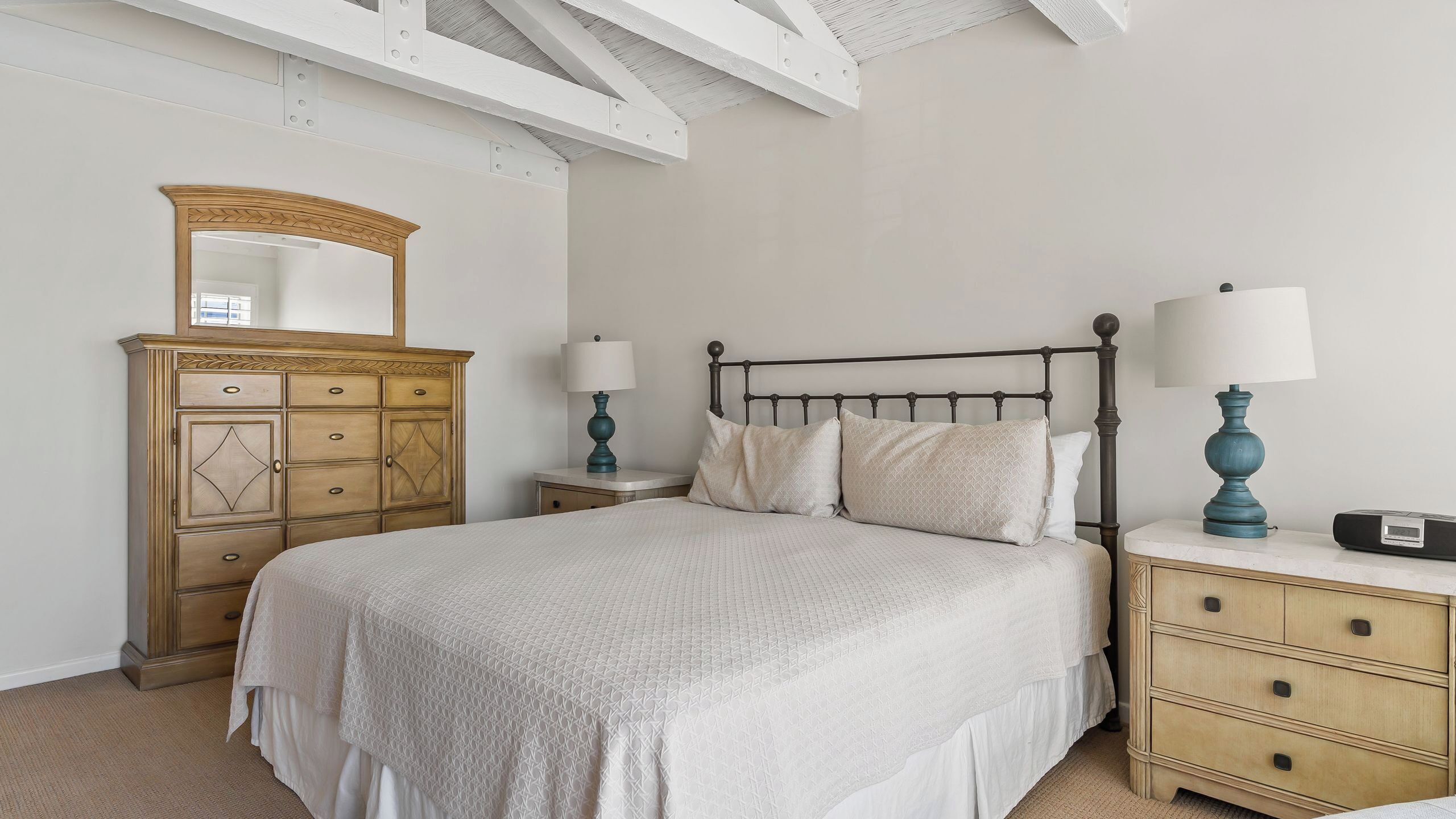 Un dormitorio con cama, mesitas de noche, cómoda, espejo y techo con vigas a la vista.