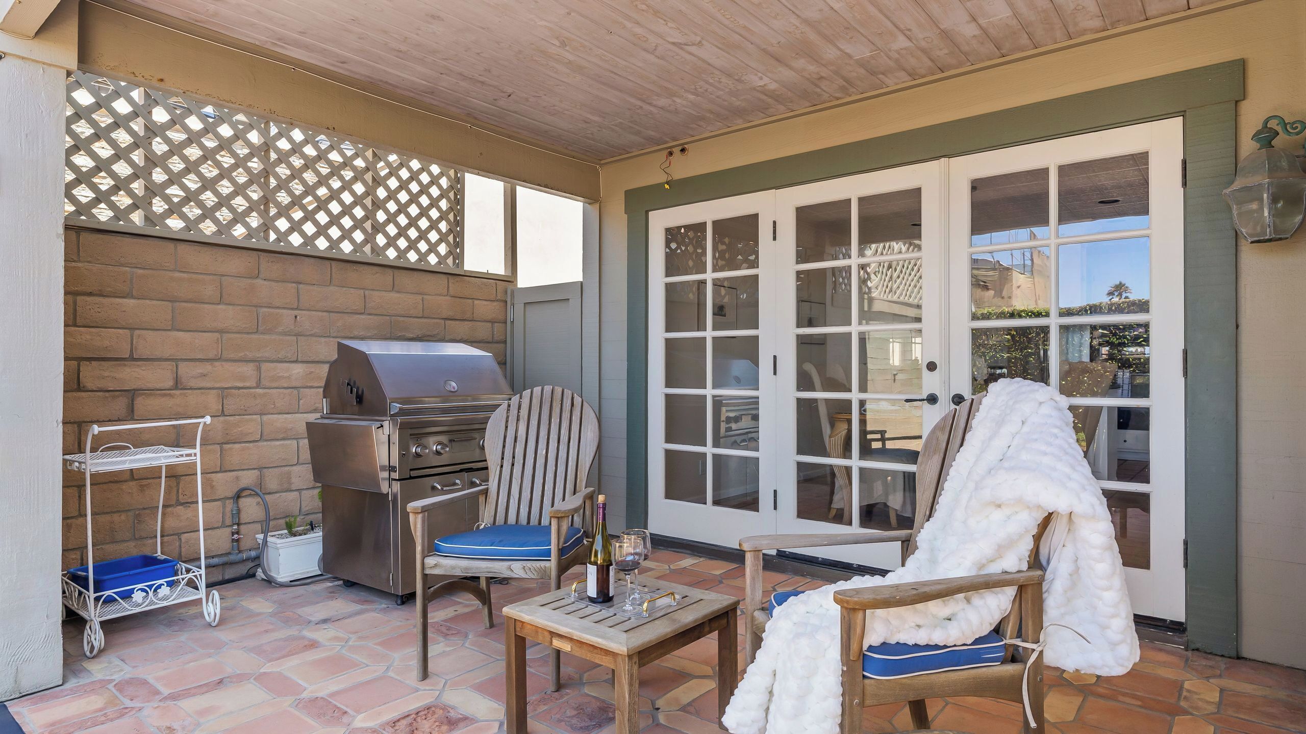 An outdoor patio with a barbecue grill, two chairs, a table with a bottle and glasses, and a sliding glass door.