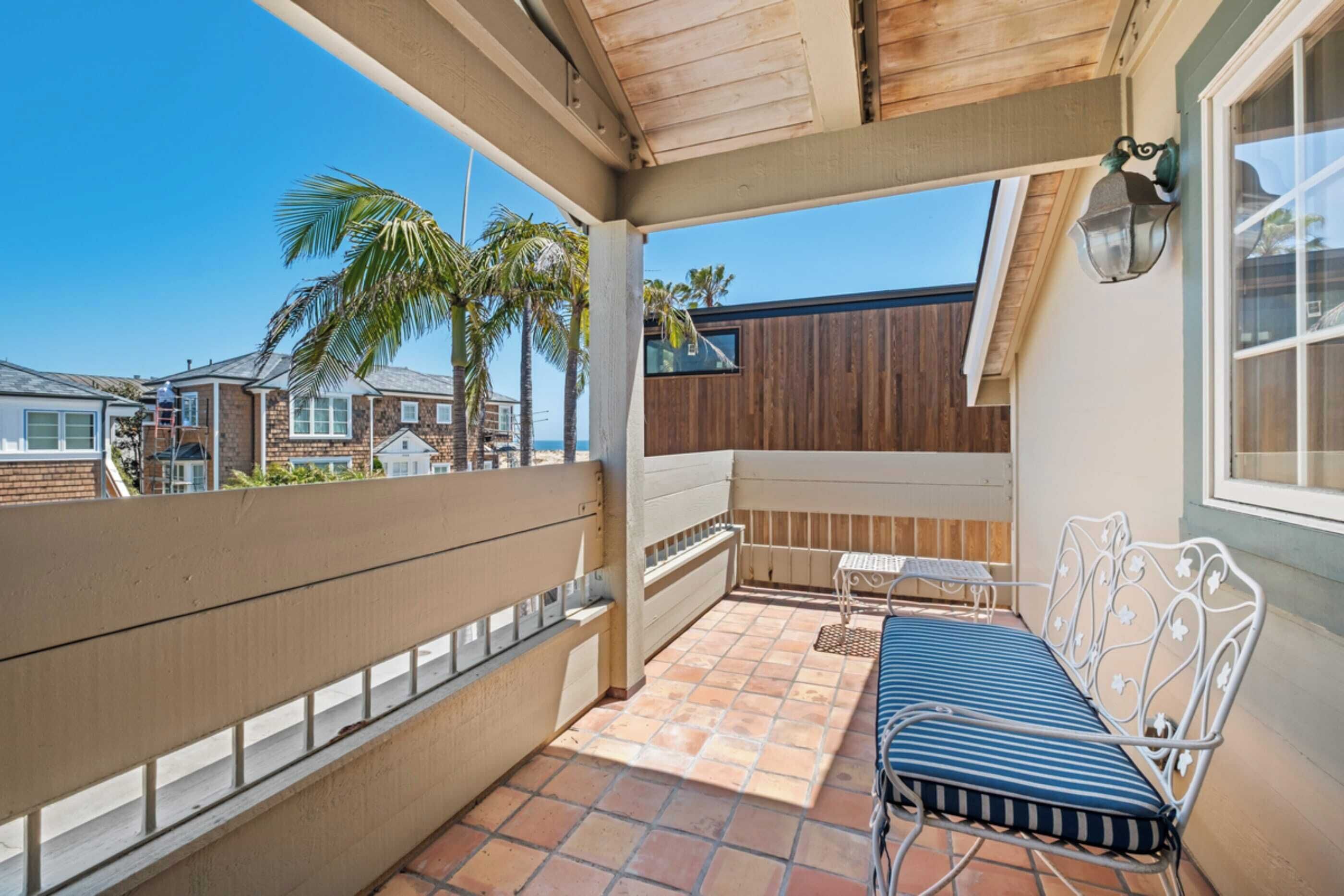 A covered balcony with a metal bench, a small table, and a view of palm trees and neighboring houses.