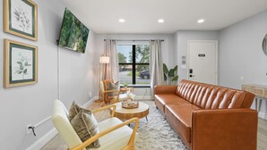 A living room with a brown leather sofa, a white armchair, a wooden coffee table, and a TV mounted on the wall. - Cozy Orlando home close to parks, dining, and downtown fun (Orlando)
