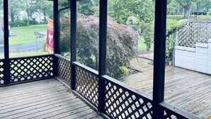 Eine überdachte Veranda mit Deckenventilator, Holzgeländer und Blick auf einen Garten mit Bäumen und Rasen.