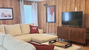 Ein Wohnzimmer mit einer Eckcouch, einem Fernseher auf einem Holzschrank und gerahmten Bildern an der Wand.