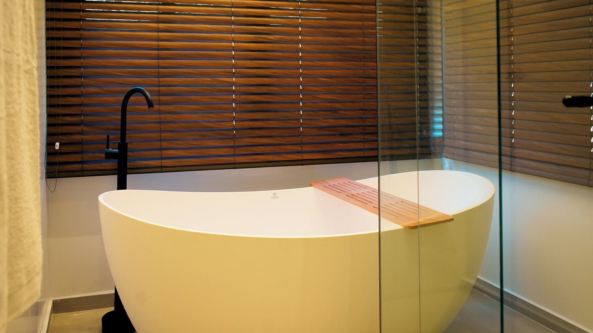 A modern bathroom with a freestanding bathtub, wooden blinds, and a glass partition.