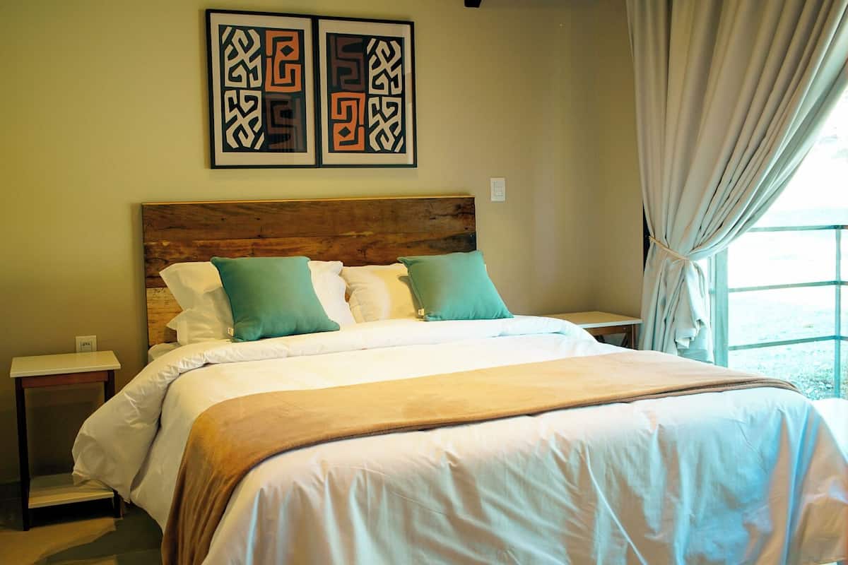 Un lit avec une tête de lit en bois, une literie blanche et des coussins verts. Deux œuvres d’art encadrées sont accrochées au-dessus du lit, et une fenêtre avec des rideaux se trouve à droite.