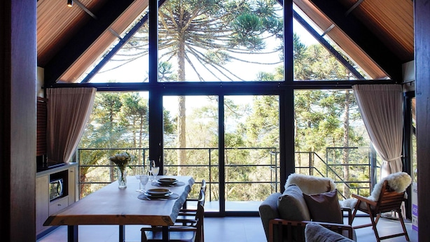 Un comedor con una mesa dispuesta para dos personas, una chimenea y un amplio ventanal con vistas a árboles.