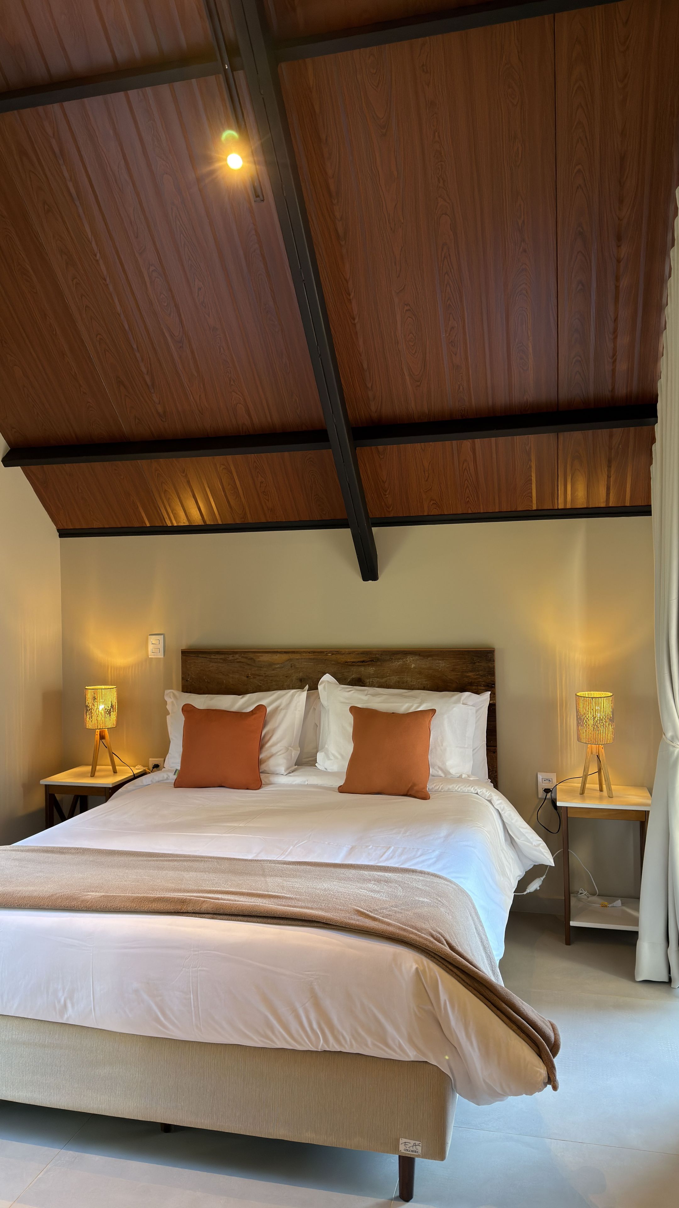 Un dormitorio con techo de madera, una cama grande con ropa de cama blanca y naranja, y dos lámparas de noche.