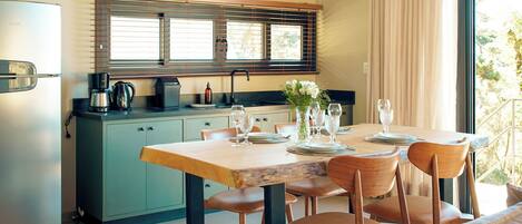 A modern kitchen with a dining area, featuring a wooden table set for four, a refrigerator, and a window with blinds.