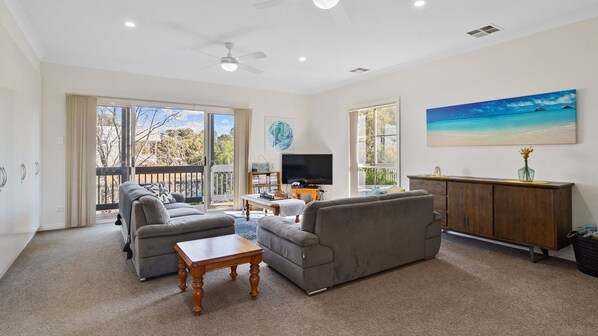 A spacious living room with a large sofa, a coffee table, and a television. There is a balcony with a view of trees and a painting of a beach on the wall.