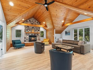 A spacious living area with a stone fireplace, wooden ceiling beams, and large windows offering views of the outdoors.