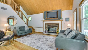 Uma sala de estar ampla com lareira, uma televisão montada acima dela e uma escada que leva a um andar superior.