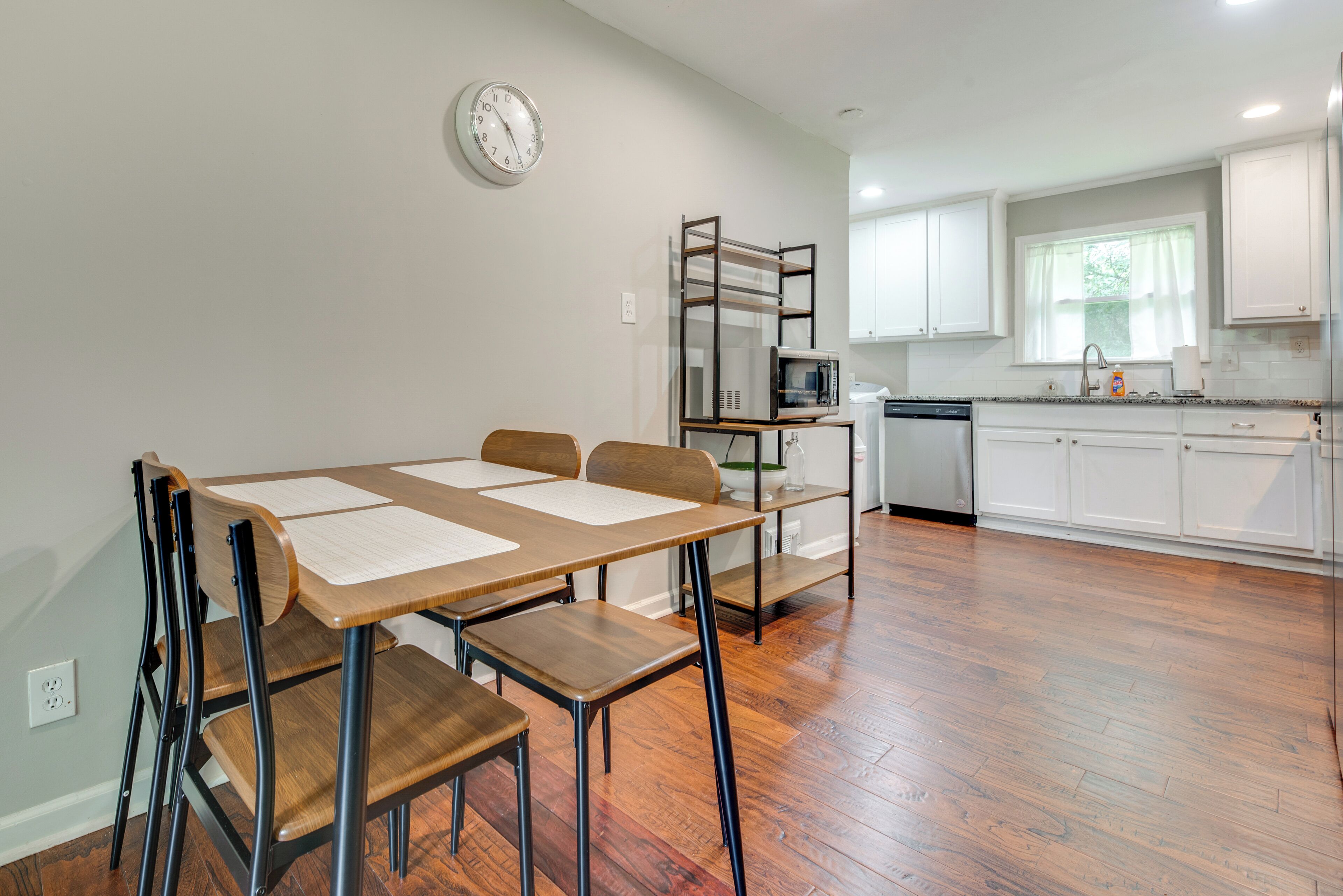Een moderne keuken met witte kasten, een magnetron en een vaatwasser. Er is een eettafel met stoelen en een klok aan de muur.