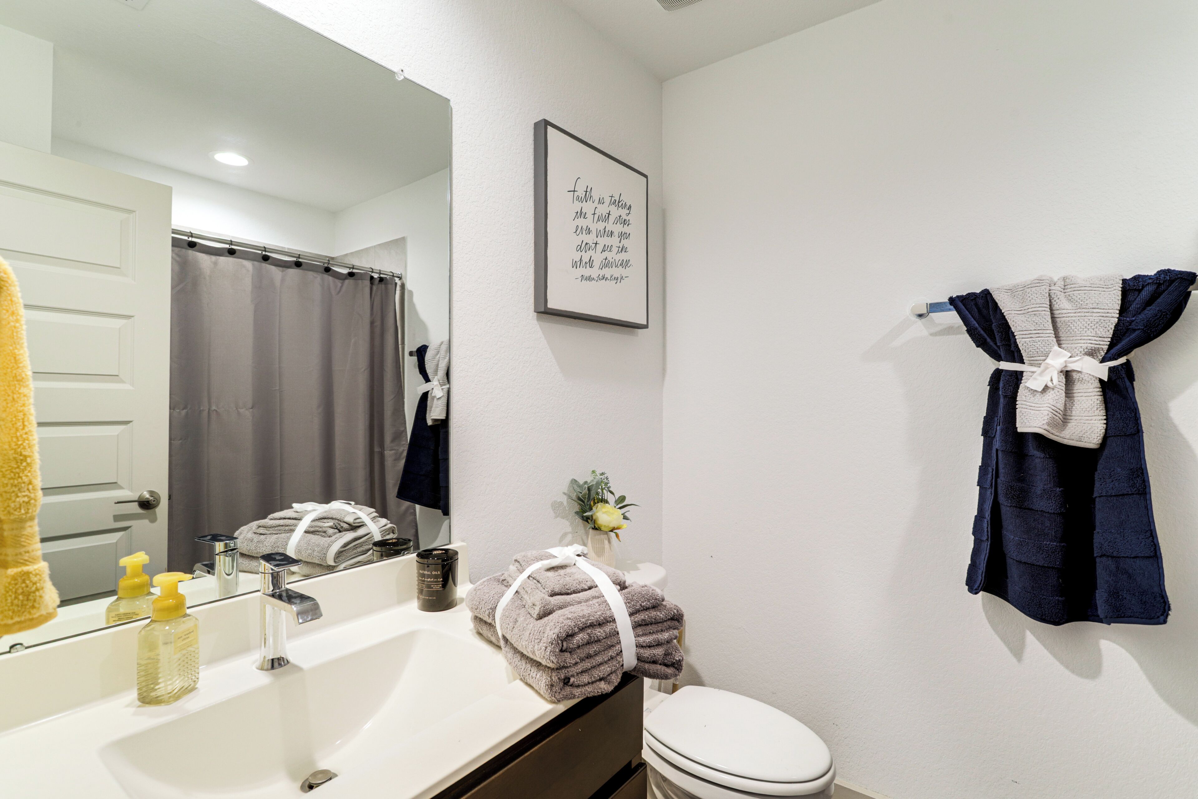 Une salle de bain avec un lavabo blanc, un miroir, une citation inspirante encadrée, des toilettes et des serviettes.