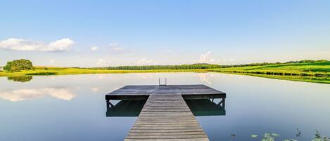 A wooden pier extending into calm water, surrounded by green fields and a clear sky.