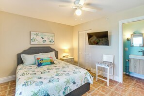 Ein Schlafzimmer mit Bett, Nachttisch, Sessel, Deckenventilator, Fernseher und einem Badezimmer mit Waschbecken und Spiegel.