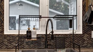 Eine Küche mit einem schwarzen Wasserhahn, einem Waschbecken und einem Fenster darüber. Auf einem Ablagerost befinden sich zwei Flaschen, auf denen „DISHES“ und „HANDS“ stehen.