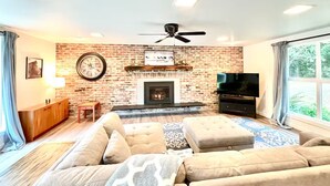 A living room with a brick fireplace, a ceiling fan, a large TV, and a sofa. - Luxury 3BR Home + Game Room - Wooded Escape Near Downtown Redmond (Redmond)