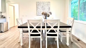 A dining area with a dark wooden table and white chairs, a decorative wall piece, and a window with a view of greenery. - Luxury 3BR Home + Game Room - Wooded Escape Near Downtown Redmond (Redmond)