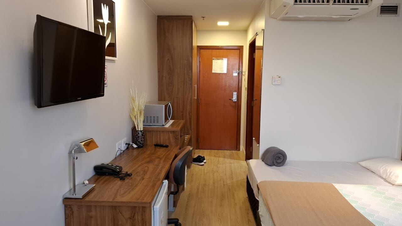 Quarto de hotel com televisão de tela plana, escrivaninha com luminária, micro-ondas, cama e porta que dá para outro cômodo.