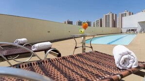 A rooftop pool area with lounge chairs, a table with drinks, and a city skyline in the background.