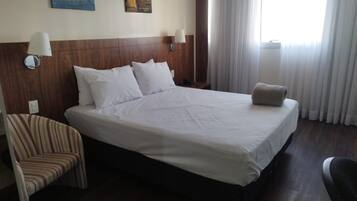 Habitación de hotel con una cama grande, dos lámparas de noche, una silla y un ventanal con cortinas.
