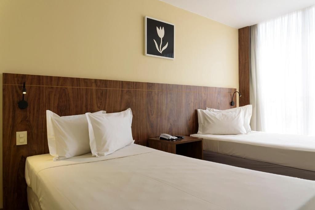 A hotel room with two beds, a wooden headboard, a wall-mounted light, and a window with curtains.