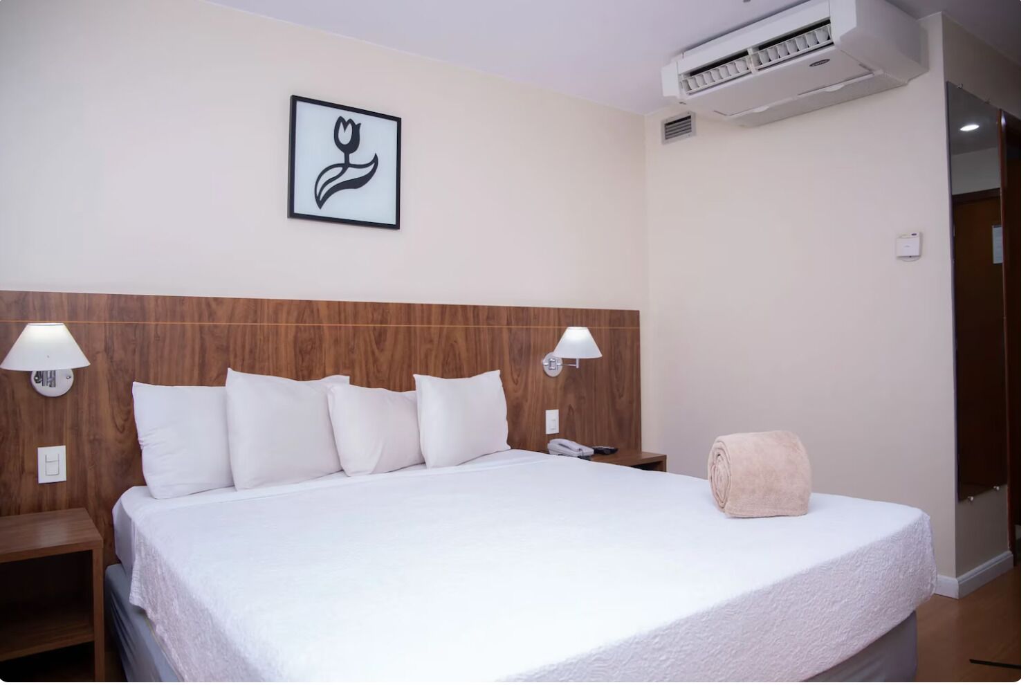 A hotel room with a wooden headboard, a bed with white bedding, two wall-mounted lamps, a framed picture, an air conditioning unit, and a rolled towel on the bed.