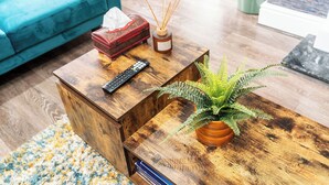 Uma mesa de centro de madeira com uma planta de samambaia, um controle remoto, uma caixa de lenços e difusores de vime numa sala de estar com um sofá azul-turquesa e um tapete estampado.