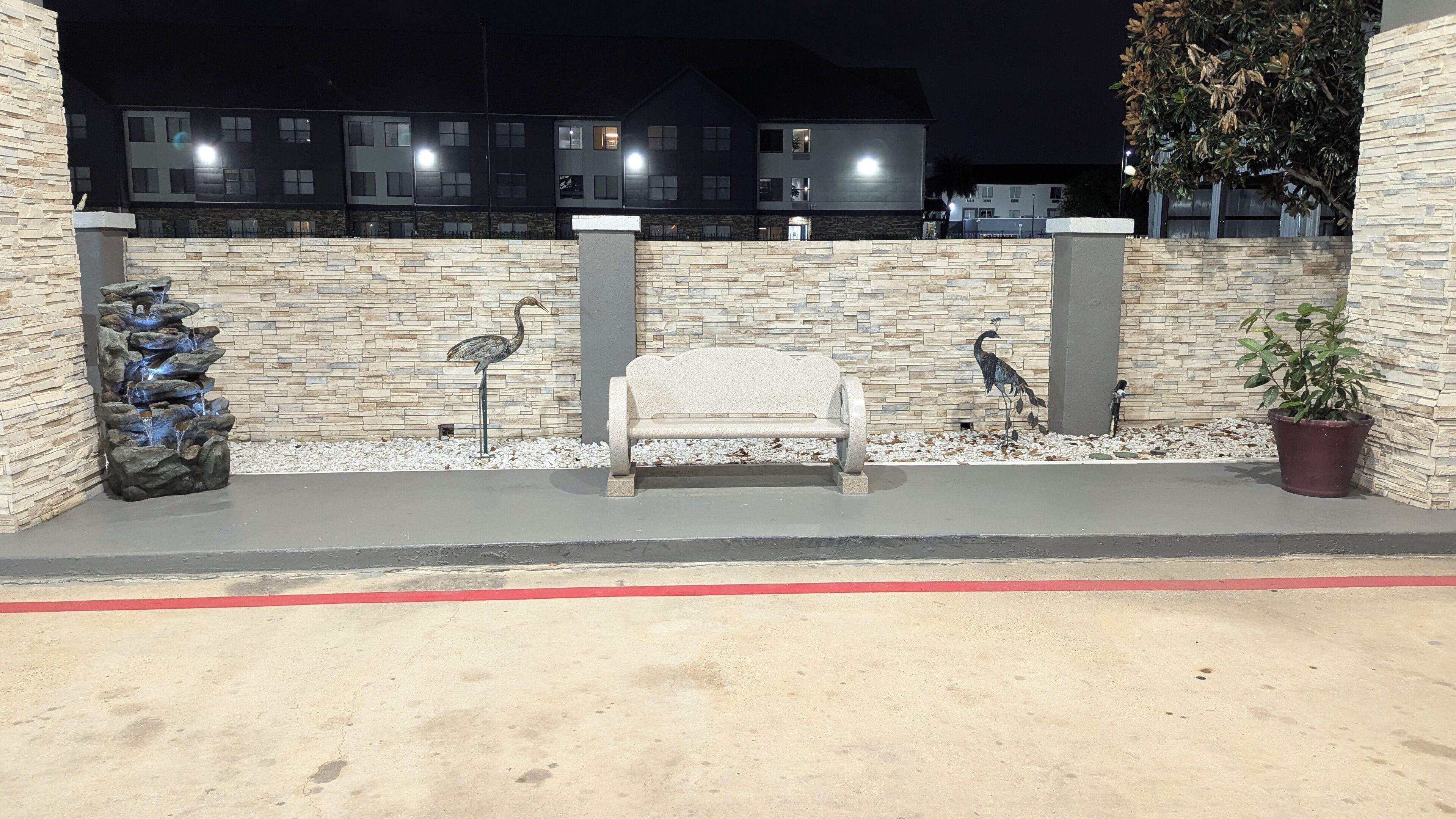 An outdoor seating area with a stone bench, a decorative fountain, and a potted plant.