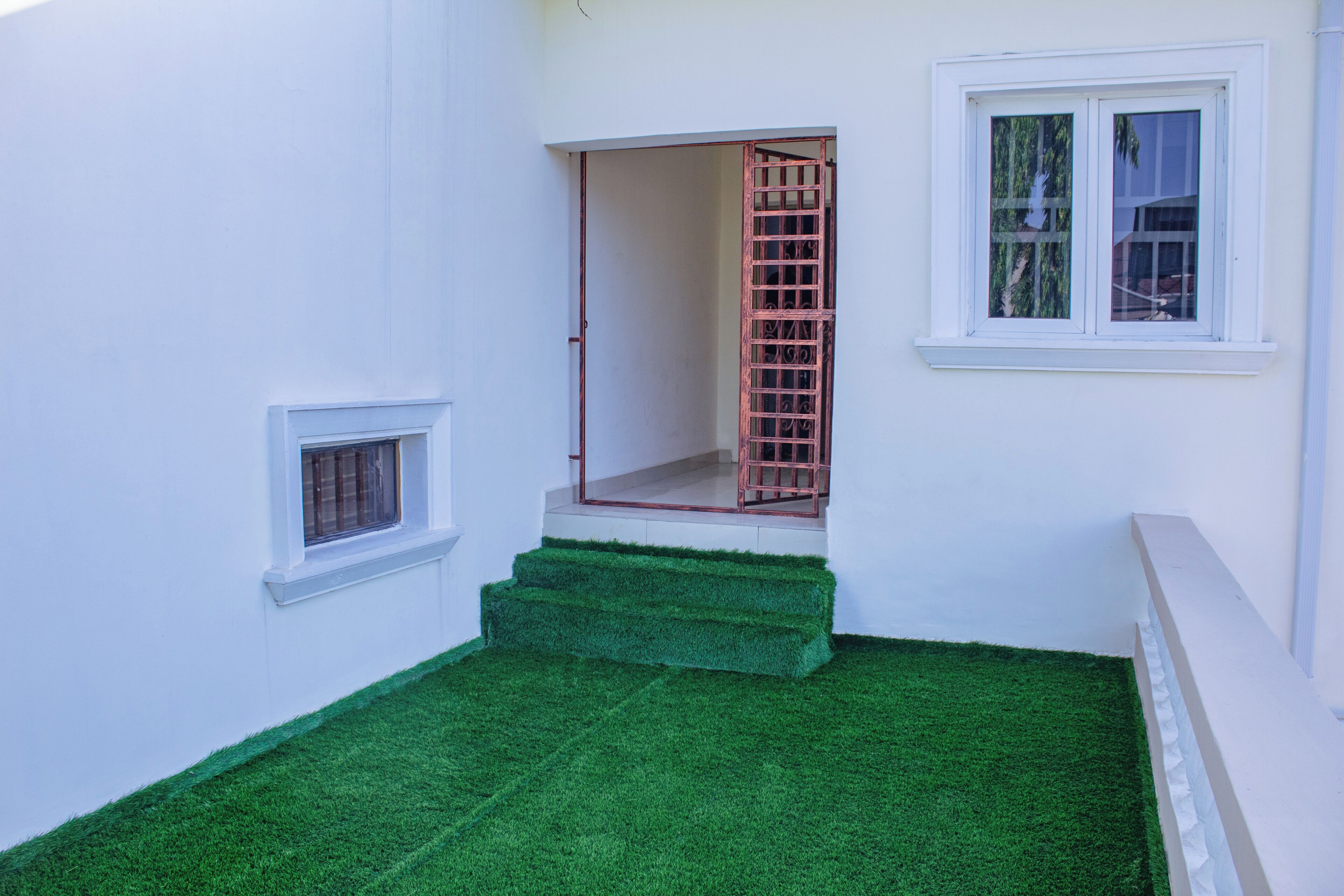 Un edificio blanco con una ventana pequeña, una puerta con reja metálica y un área de césped artificial verde.