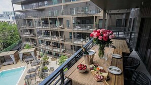 Un balcón con piscina, una mesa de comedor con comida y bebidas, y vistas de un edificio de varios pisos.