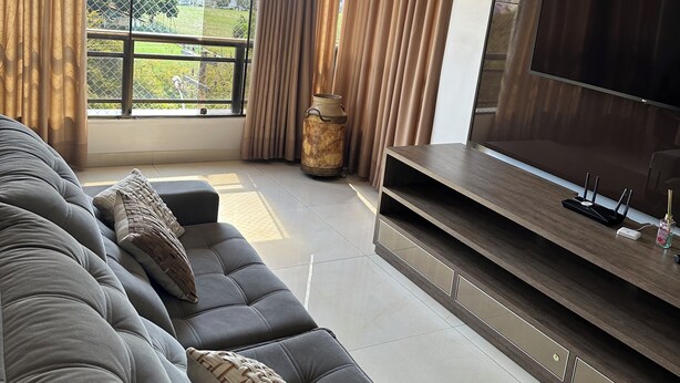 A modern living room with a large grey sofa, a flat-screen TV, and a view of a park and buildings through large windows. - Make yourself at home (Goiania)